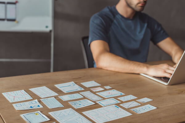 A developer typing on a silver laptop at a wooden desk with various paper cut-out UI wireframe components arranged in the foreground.