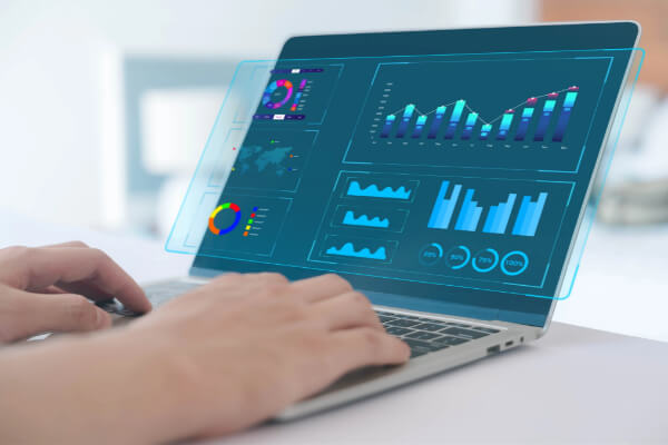 Hands typing on a silver laptop keyboard while a glowing blue, futuristic holographic data dashboard projects upward from the screen.