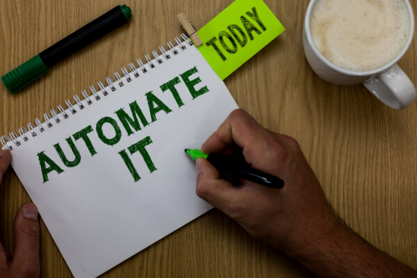 Top-down view of hands using a marker to write AUTOMATE IT on a white spiral notebook, next to a green sticky note reading TODAY and a coffee cup.