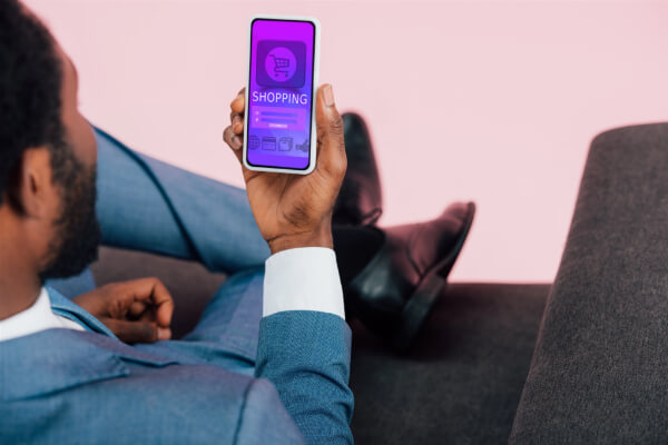 A person in a blue suit sitting down while holding a smartphone that displays a purple mobile shopping app with a cart icon.