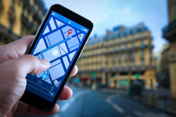 A hand holding a smartphone displaying a digital GPS street map with a blue route line and a red location pin, with a blurred city street in the background.