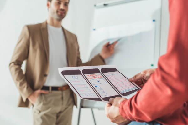 A close-up of a person holding printed mobile phone wireframes, with a man in a tan blazer presenting at a whiteboard in the background.