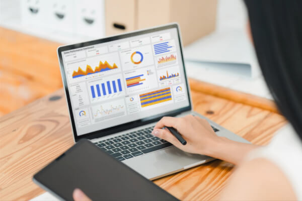 Over-the-shoulder view of a woman using a stylus and looking at a laptop screen that displays a bright sales performance dashboard with colorful charts.